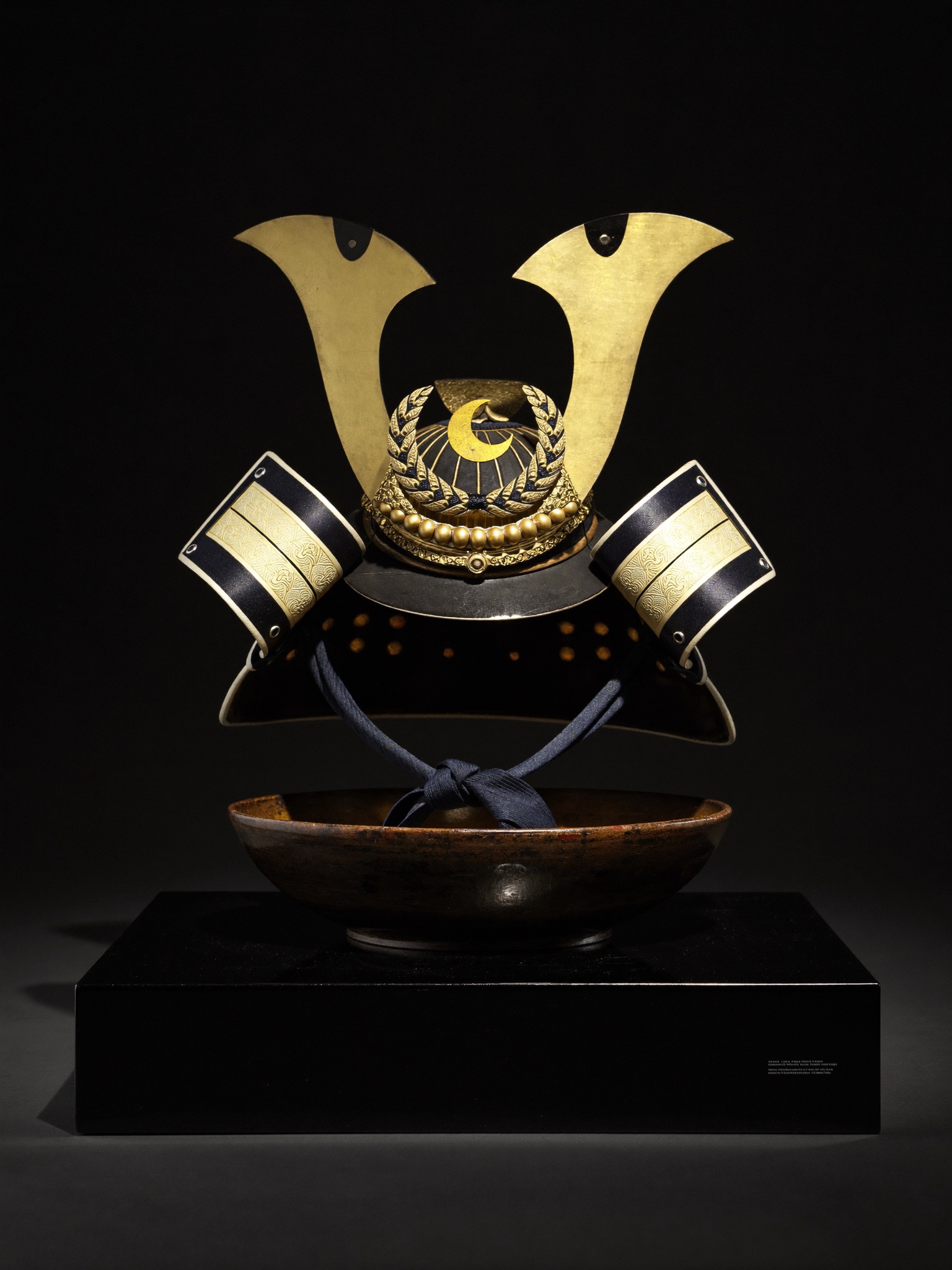 Lacquered kabuto helmet with a gold crescent-moon crest, lit by a single warm light against darkness
