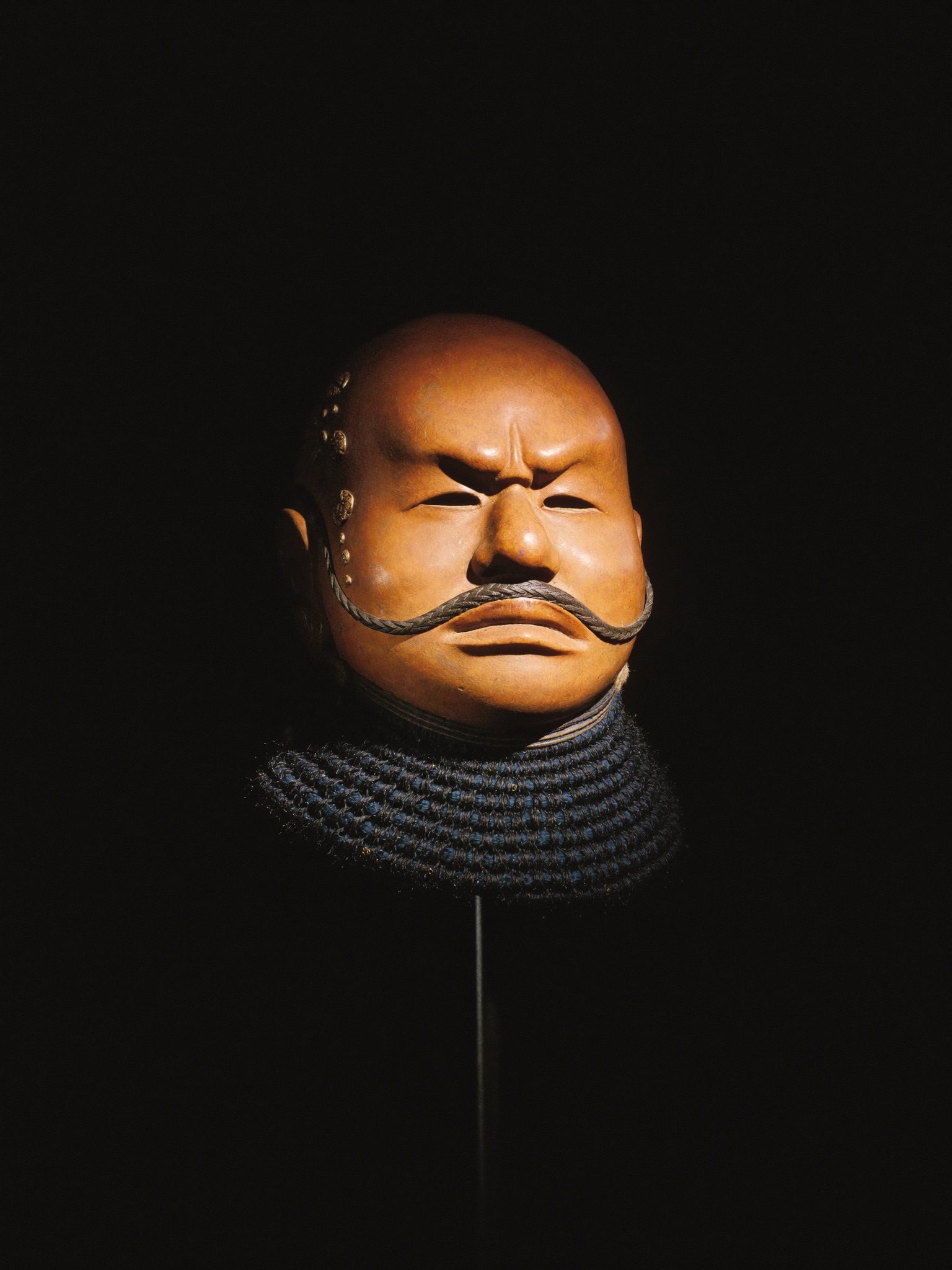 Lifelike russet-lacquered full face mask with gold repair beads above the left brow, mounted in darkness